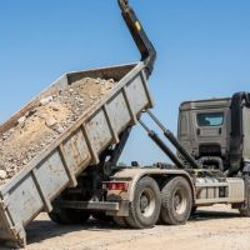 Camión volquete gris descargando escombros en un terreno de construcción bajo un cielo azul.